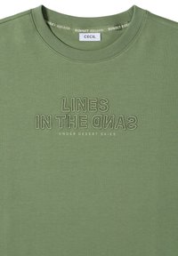 T-shirt vert col rond avec texte brodé "LINES IN THE SAND" et texte imprimé plus petit "UNDER DESERT SKIES" sur le devant.