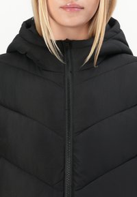 Zwarte pufferjack met een chevron-quiltontwerp, hoge kraag en een sluiting met een rits aan de voorkant. Het materiaal lijkt soepel en geïsoleerd te zijn.