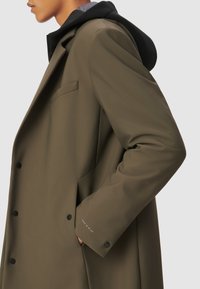 Persona con un abrigo de doble botonadura verde oliva y botones negros, con una sudadera negra debajo, mano en el bolsillo del abrigo, perfil lateral.