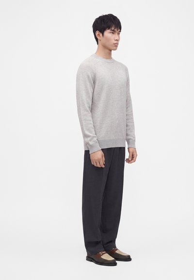 Homme de profil portant un pull à manches longues gris clair, un pantalon noir ample et des mocassins marron et beige sur fond blanc.