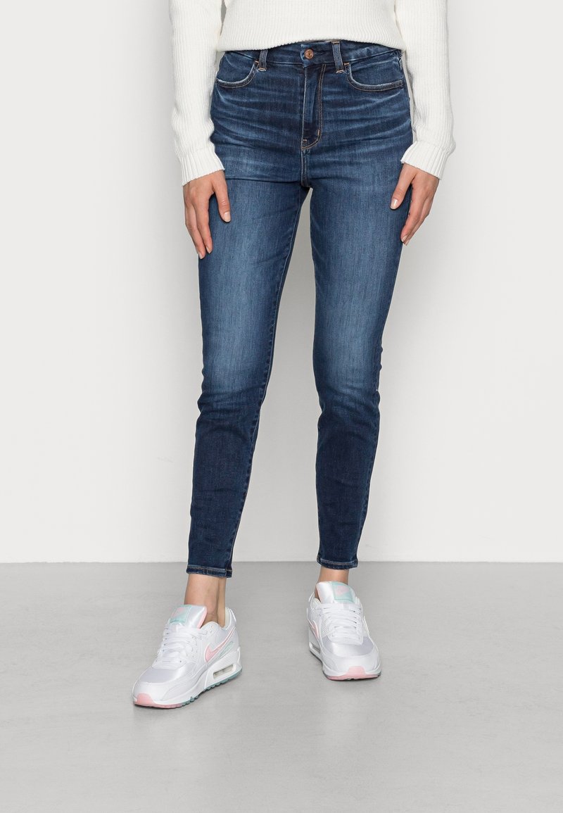 Personne portant un jean slim bleu, un pull blanc et des baskets blanches avec des accents rose et menthe, debout sur un sol gris devant un mur uni.