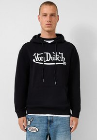 Zwarte hoodie met voorzak, witte "Von Dutch" logo en trekkoorden. Gecombineerd met blauwe jeans met logo patches.