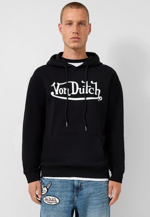Czarna bluza z kapturem, z przednią kieszenią, białym logo "Von Dutch" oraz sznurkami. W zestawie z niebieskimi dżinsami z łatami z logo.