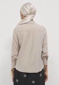 Camicia lunga beige chiaro a maniche lunghe con tessuto testurizzato, un plissè sul retro e polsini abbottonati, abbinata a una sciarpa liscia e lucida di colore chiaro e una gonna ricamata con fiori.