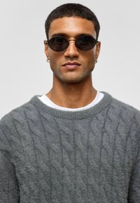 Grijze gebreide trui met een kabelpatroon, gedragen over een witte T-shirt. Zwarte ovale zonnebril met dunne monturen complementeren de outfit.