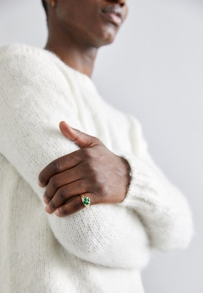 Goldring mit grünen Akzenten, platziert auf einem strukturierten weißen Strickpullover. Die Hand posiert anmutig auf dem Arm.