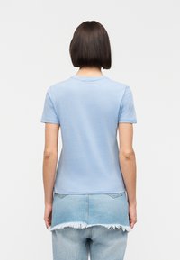 Personne aux cheveux courts et foncés portant un t-shirt bleu clair et une jupe en denim délavé à plusieurs couches par-dessus un jean, vue de dos.