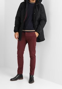 Uomo che indossa un parka nero, un maglione blu navy con colletto a righe, pantaloni bordeaux e stivali alla caviglia in pelle nera, in piedi contro un muro bianco.