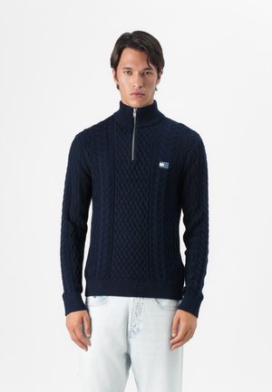 Marineblauwe gebreide trui met hoge kraag, voorzien van kabelpatronen en een kwart-rits. Geribbelde manchetten en zoom, met een klein logodetail.