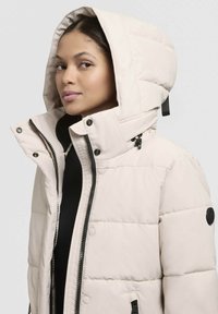 Helle beige Daunenjacke mit großer Kapuze, Reißverschluss- und Knopfverschluss vorne, schwarzen Akzenten und gestepptem Muster. Verfügt über elastische Handgelenkabschlüsse.
