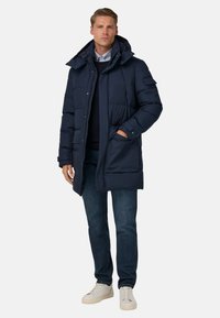 Námořnická péřová bunda s kapucí, zipovým uzávěrem a předními kapsami; spárována s tmavými džíny a bílými teniskami s texturovaným povrchem.