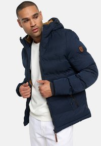 Marinblå pufferjacka med huva, med brunt foder och sidofickor. Inkluderar dragkedja och läderdetaljer. Mjuk textur.