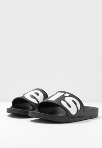 Sandales noires à glissière avec de larges brides arborant un grand texte blanc, une semelle plate et une semelle intérieure texturée pour une meilleure adhérence.