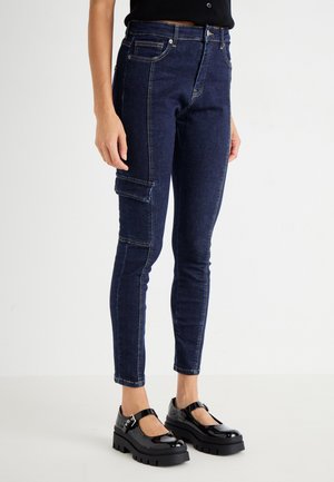 Slanke cargo jeans in donkerdenim met een zijzak, contrasterende stiksels en een getailleerd ontwerp. Gecombineerd met stevige zwarte platform schoenen.