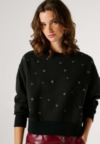 Melns džemperis ar sudraba acs detaļām, apaļu apkakli un ribotām aprocēm. Mīksts audums ar brīvu piegriezumu un bez redzama logotipa.