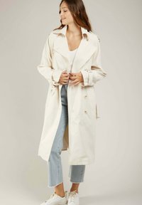 Trench-coat beige clair avec un design à double boutonnage, un grand col et des poignets ceinturés, associé à un jean cropped bleu clair.