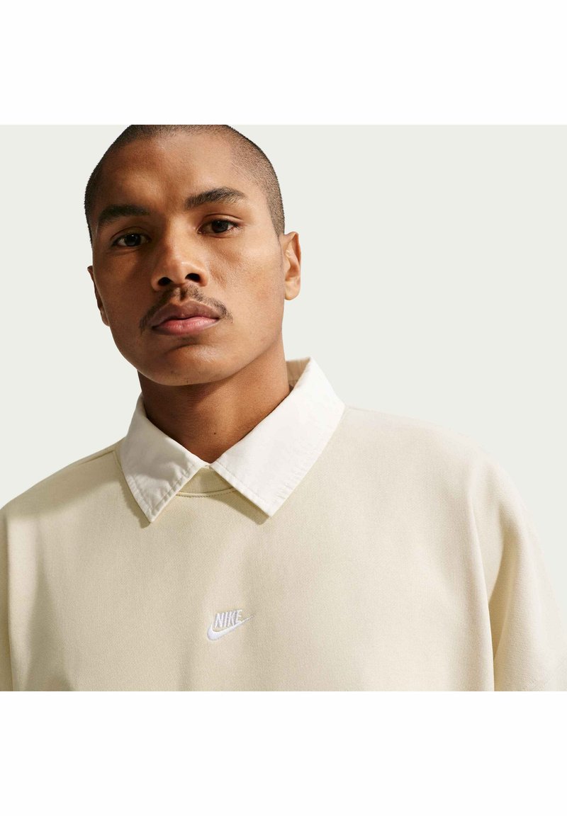 Homme aux cheveux courts et à la moustache porte un pull Nike crème avec un col blanc, face à un fond clair uni.