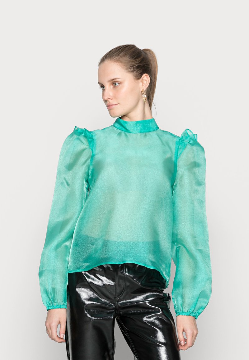 Blusa transparente em turquesa com mangas longas e bufantes, com detalhes em babados nos ombros. Combinada com calças pretas brilhantes.