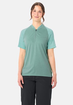 Giovane donna con una maglietta teal a maniche corte con cerniera e pantaloni neri, in piedi davanti a uno sfondo chiaro uniforme, che sorride dolcemente.