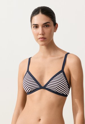 Junge Frau mit dunklen, zurückgebundenen Haaren, die ein dreieckiges Bikinioberteil mit marineblauen und weißen Streifen vor einem einfarbigen Hintergrund trägt.