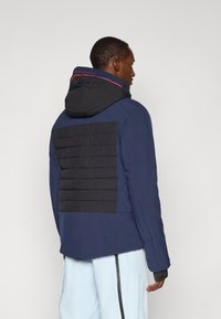 Marineblaues Jacket mit einem schwarzen, steppbaren Rückenteil, Kapuze und kontrastierenden roten Akzenten am Kragen. Entwickelt für Wärme und Funktionalität.