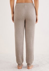 Femme debout pieds nus sur un tapis, portant un pantalon de détente en tricot beige, vue de dos sur un fond uni.