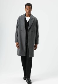 Grijze wollen overjas met een brede kraag, dubbele rij knopen en een losse pasvorm, gedragen over een zwart vest en zwarte broek. Glad stoffen textuur.