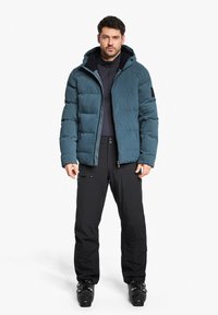 Padded tealfarbige Jacke mit Kapuze, ausgestattet mit einem Reißverschluss und seitlichen Taschen, kombiniert mit schwarzen Hosen und Stiefeln. Glatte Textur, warmes Design.