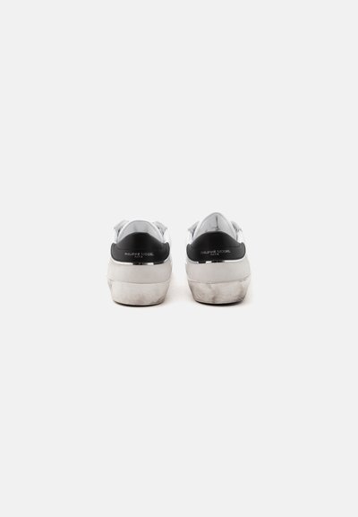 Paire de baskets blanches avec des languettes noires au talon affichant les logos "Philippe Model Paris", vue de dos sur fond blanc.