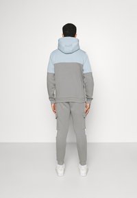 Grauer und hellblauer Kapuzenpullover mit Kängurutasche, kombiniert mit grauer Cargohose mit Seitentaschen. Weiße Turnschuhe.