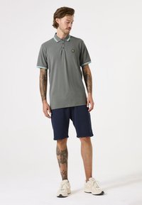 Grijze poloshirt met een twee-knopen placket en een kraag met turquoise strepen, gecombineerd met marineblauwe shorts en crèmekleurige sneakers. Bevat zichtbare tatoeages.