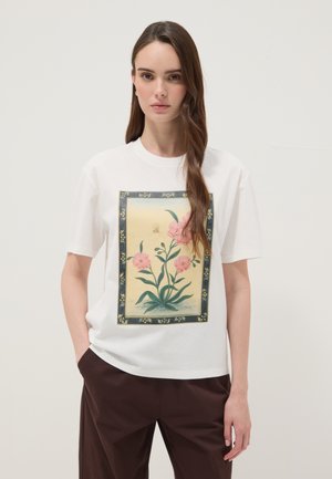 Frau mit langen braunen Haaren, die ein weißes T-Shirt mit einem gerahmten rosa Blumen- und Schmetterlingsdruck trägt, kombiniert mit dunkelbraunen Hosen.