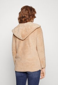 Femme aux cheveux roux bouclés portant une veste polaire beige à capuche et un jean bleu, vue de dos sur un fond uni.