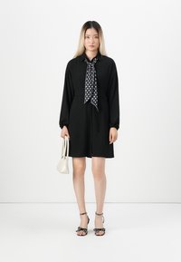 Black dress with long sleeves and waist tie, featuring a polka dot scarf. White handbag and black heeled sandals with bow accents complement the outfit.