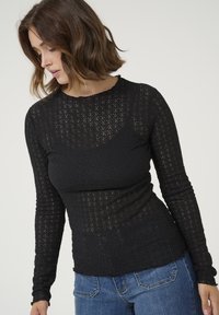 Haut noir à manches longues en tissu semblable à de la dentelle avec un motif texturé. Design ajusté avec un col rond, associé à un jean en denim bleu.