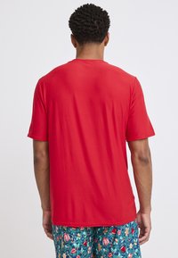 Czerwona koszulka z krótkim rękawem i okrągłym dekoltem, o gładkiej fakturze i luźnym kroju, noszona z szortami w wzory z motywami wakacyjnymi.