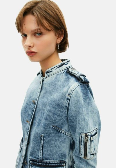 Veste en denim bleu clair avec un col, plusieurs poches et une poche latérale zippée. Présente une texture décolorée et des fermetures à boutons.