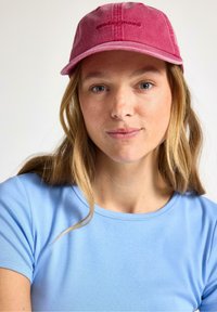 Roze katoenen pet met geborduurde tekst "zondagstemming"; heeft een stevige rand en een verstelbare achterkant. Draagt bij een lichtblauw shirt.