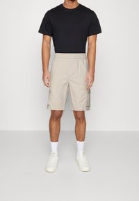 Hombre vestido con una camiseta negra, pantalones cortos beige tipo cargo, calcetines blancos y zapatillas blancas, de pie frente a un fondo claro y liso.