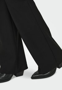 Pantaloni neri a gamba larga abbinati a scarpe nere con tacco, con punta affilata e cuciture decorative lungo i bordi superiori.