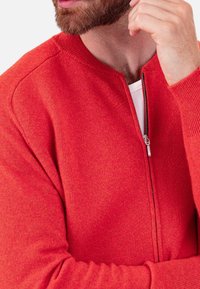 Roter Cardigan mit Reißverschluss aus weichem Strickmaterial. Verfügt über einen runden Ausschnitt, gerippte Bündchen und Reißverschluss-Details.