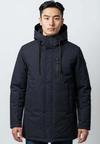 Homme portant un manteau d'hiver noir à capuche avec fermeture éclair et boutons, se tenant devant un fond clair.