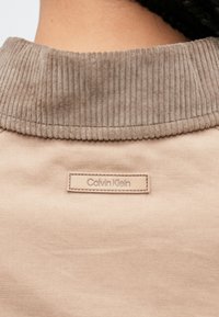 Šviesiai smėlio spalvos velveto apykaklė ant smėlio spalvos audinio drabužio su siuvinėta Calvin Klein logotipo etikete. Lygus tekstūra su juostelėmis.