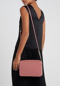 Różowa skórzana torebka crossbody o prostokątnym kształcie, ze złotym łańcuszkiem i wytłoczonym logo. Noszona na czarnej, plisowanej sukience.