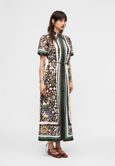 Γυναίκα που στέκεται με φόρεμα μάξι με σχέδια, κοντά μανίκια, γιακά, κάθετες ρίγες και καφέ παπούτσια με κορδόνια, μπροστά σε απλό φόντο.