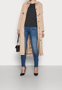 Béžový trenčkot s knoflíky, viditelná vnitřní podšívka, v kombinaci s tmavým svetrem a modrými džíny. Černé lodičky na podpatku a malá kabelka.