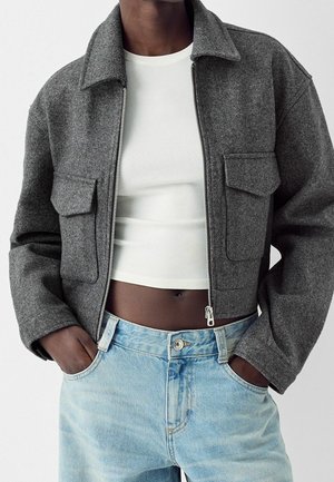 Personne portant une veste grise zippée avec de grandes poches à rabat, un top blanc court et un jean taille haute bleu clair, les mains dans les poches.