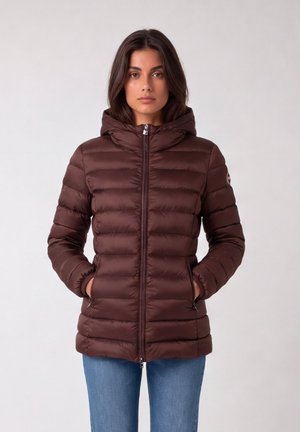 Donna che indossa una giacca trapuntata marrone con cappuccio e mani nelle tasche, abbinata a jeans blu, in piedi davanti a uno sfondo semplice.