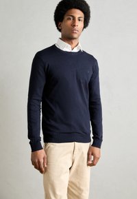 Navy blue knitted sweater with a round neck and ribbed cuffs, paired with light beige pants. Subtle logo detail on the left chest.