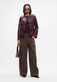 Burgundská kožená bunda s okrúhlym výstrihom, zapínaním na gombíky a prednou vreckom, doplnená hnedými širokými nohavicami a zladenou kabelkou.